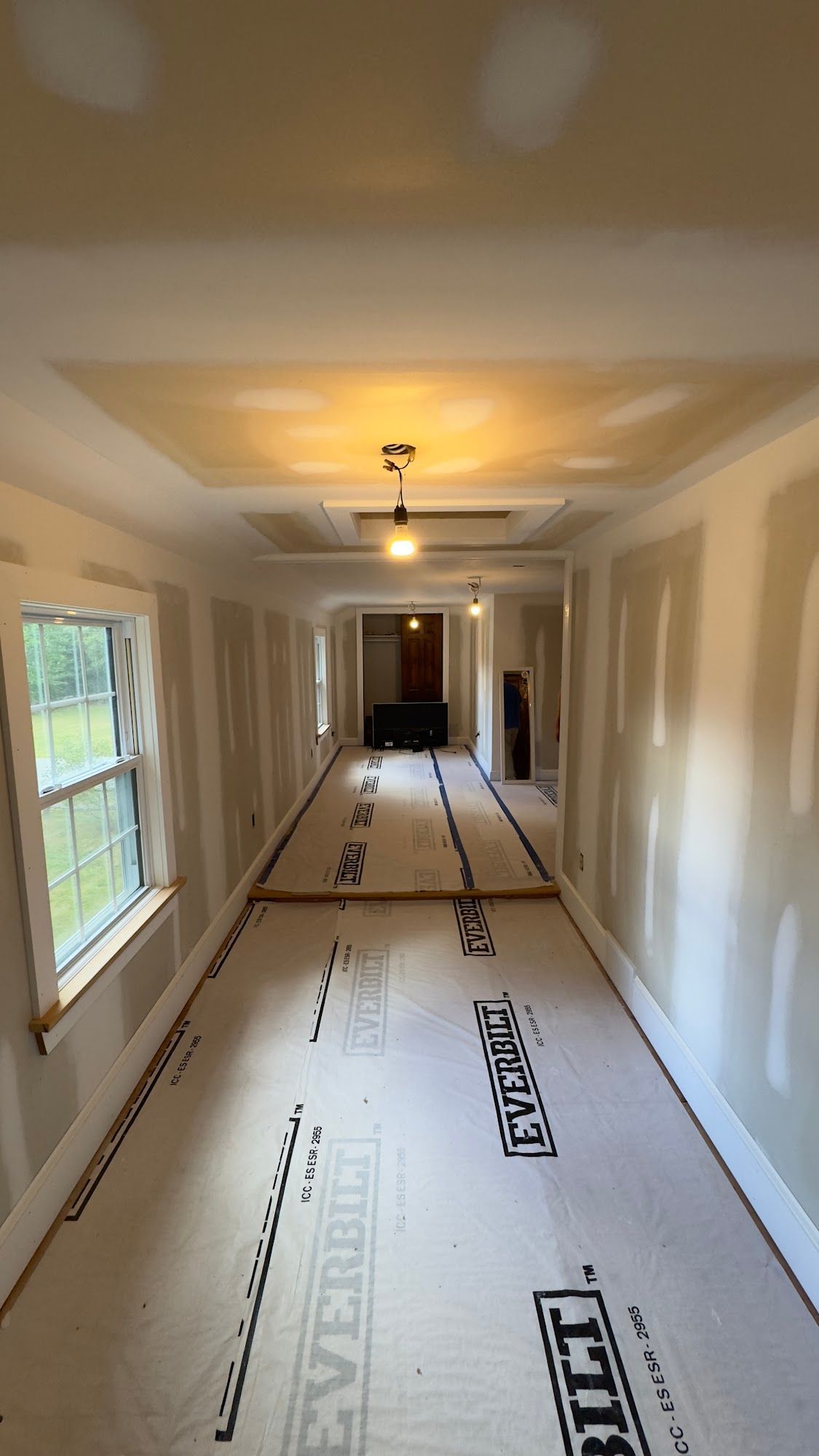 Long, unfinished hallway with drywall, protective flooring, and recessed ceiling lights.