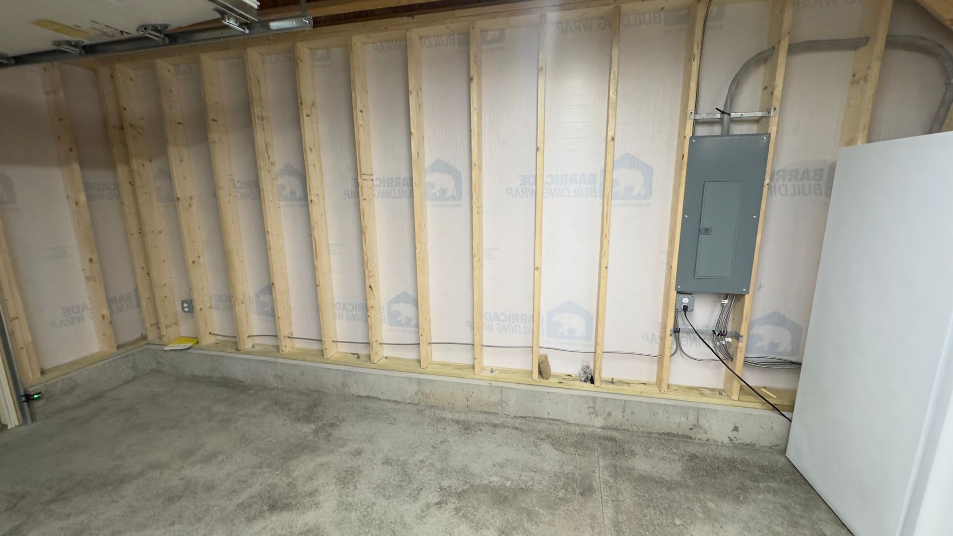 Interior view of a partially framed wall in a basement, showing studs, insulation, and electrical panel.