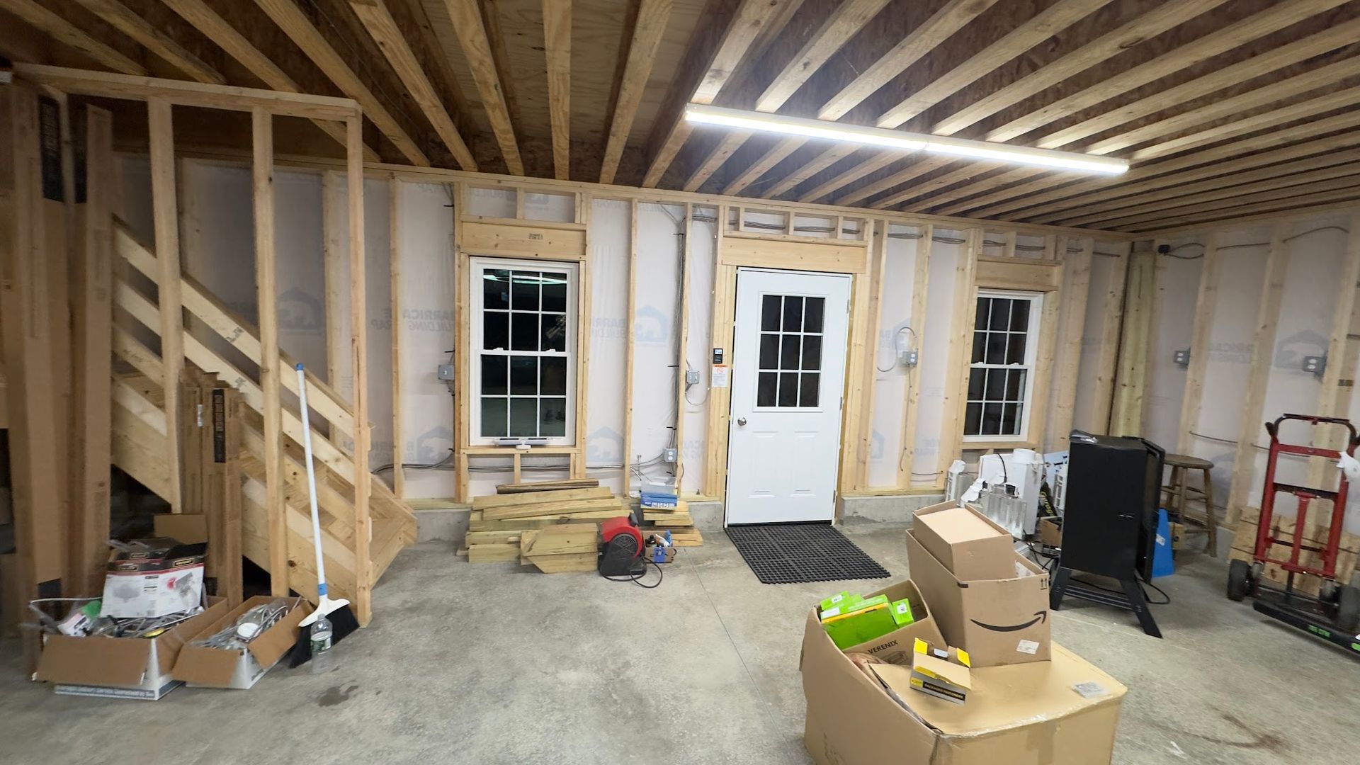 Interior of unfinished building with wood framing, windows, and door. Boxes and tools on floor.