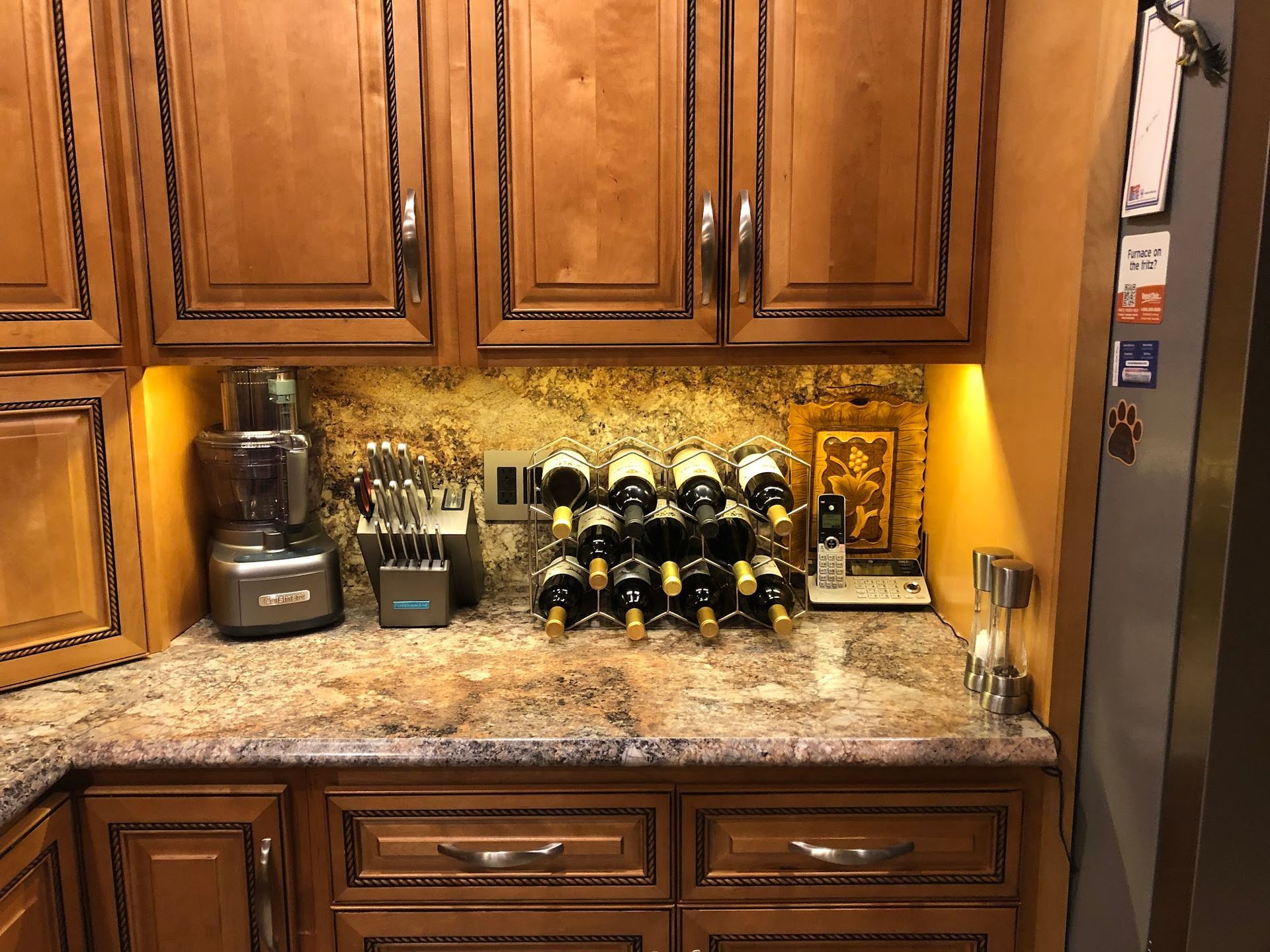 Kitchen counter with wine rack, food processor, and knife block. Brown cabinets, granite countertop, and gold backsplash.