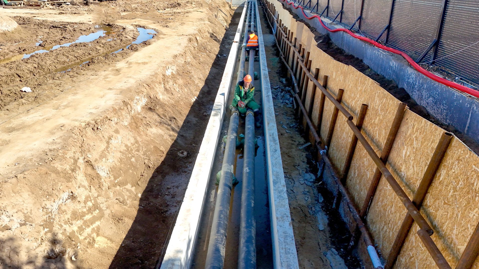 Workers inspect a deep trench with pipes beside an earthen embankment at a construction site