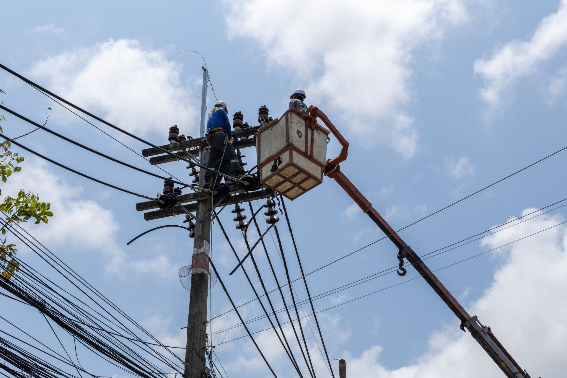 Utility workers in a bucket lift repairing power lines on a utility pole under a blue sky
