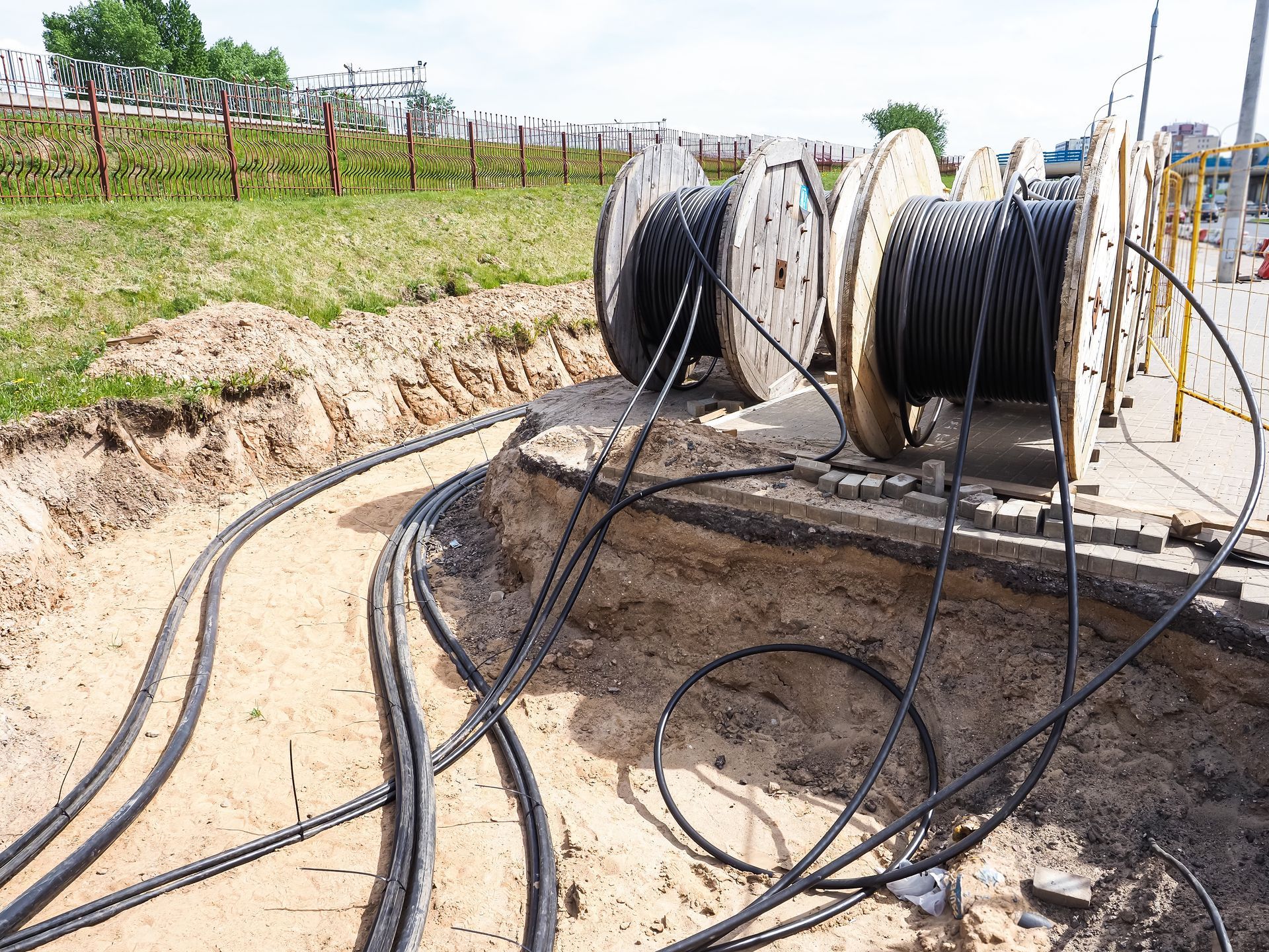 Coiled black cables on reels at a roadside trench beside a fenced grassy field
