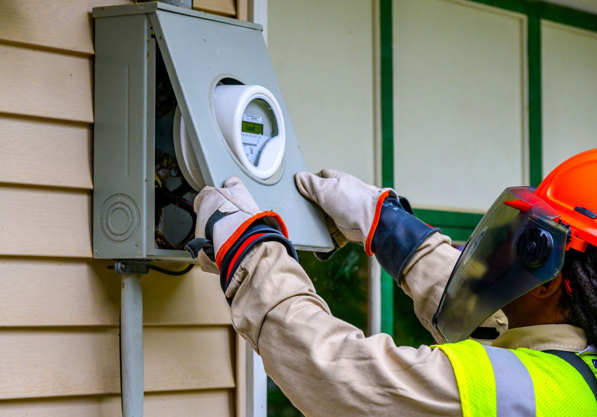 Electrician installing a utility meter on a house exterior, wearing gloves and a face shield.