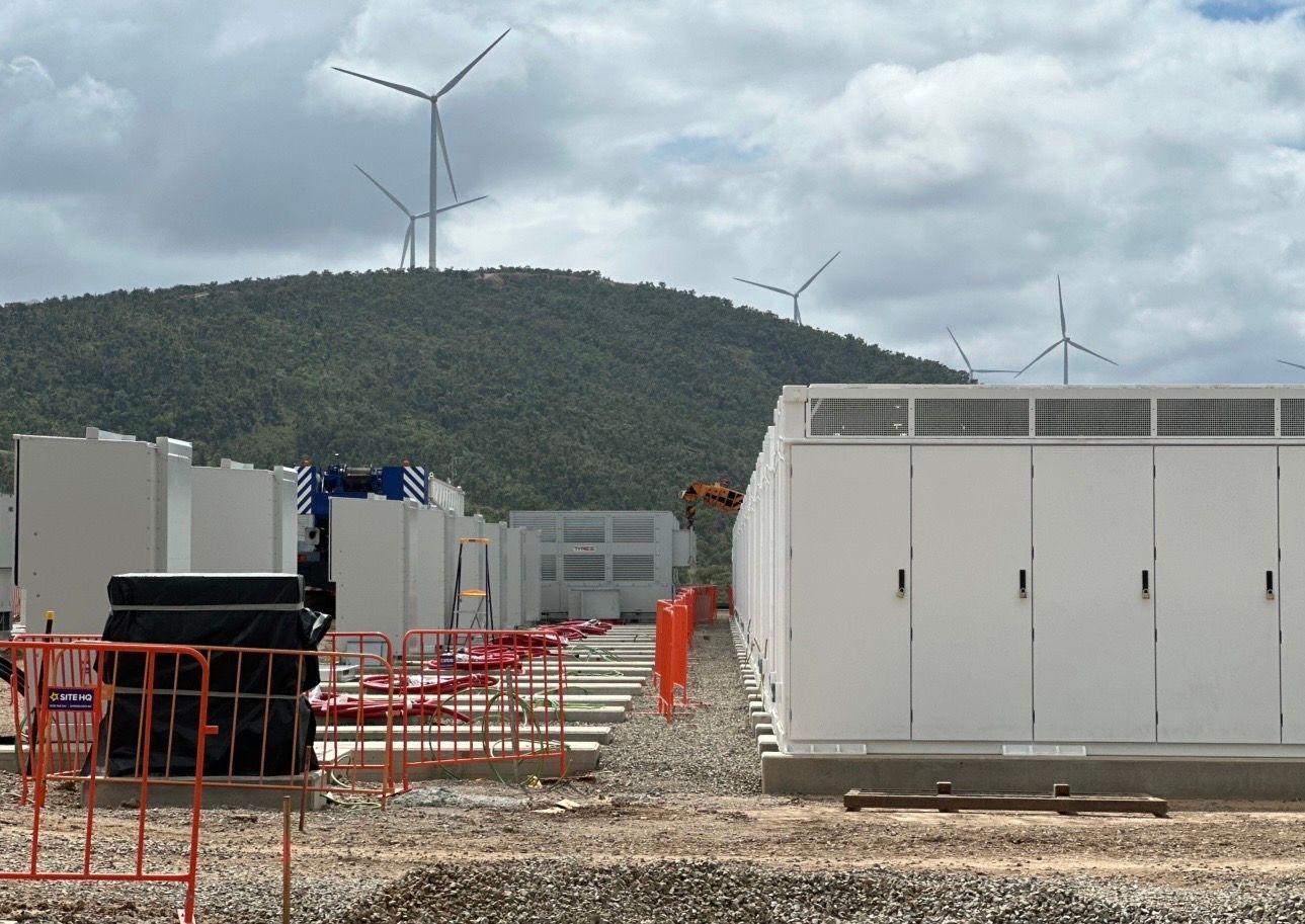 Wind turbines on a green hill behind white utility containers at a construction site.