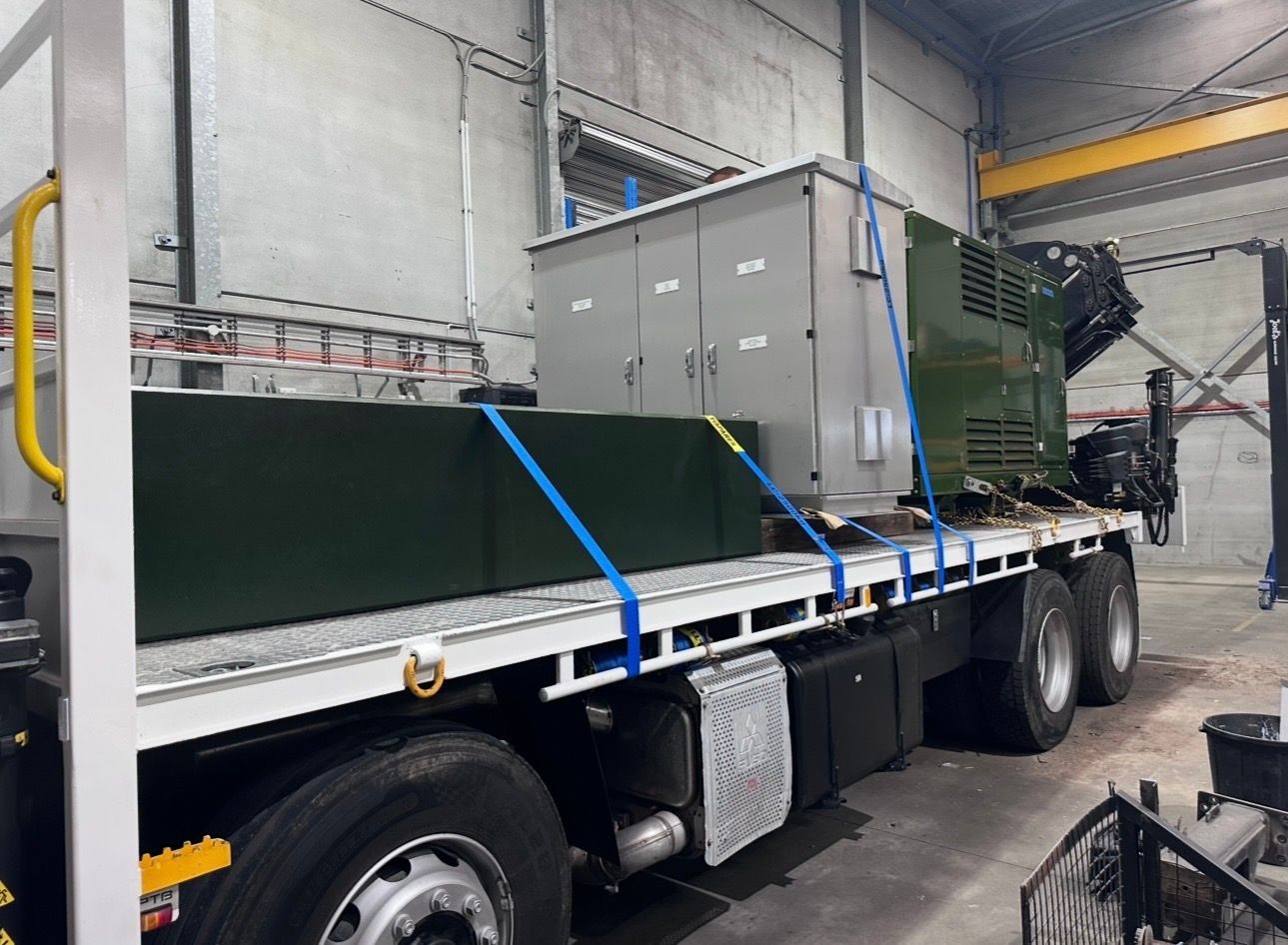 Loaded flatbed truck inside a warehouse with a large green industrial machine strapped down.