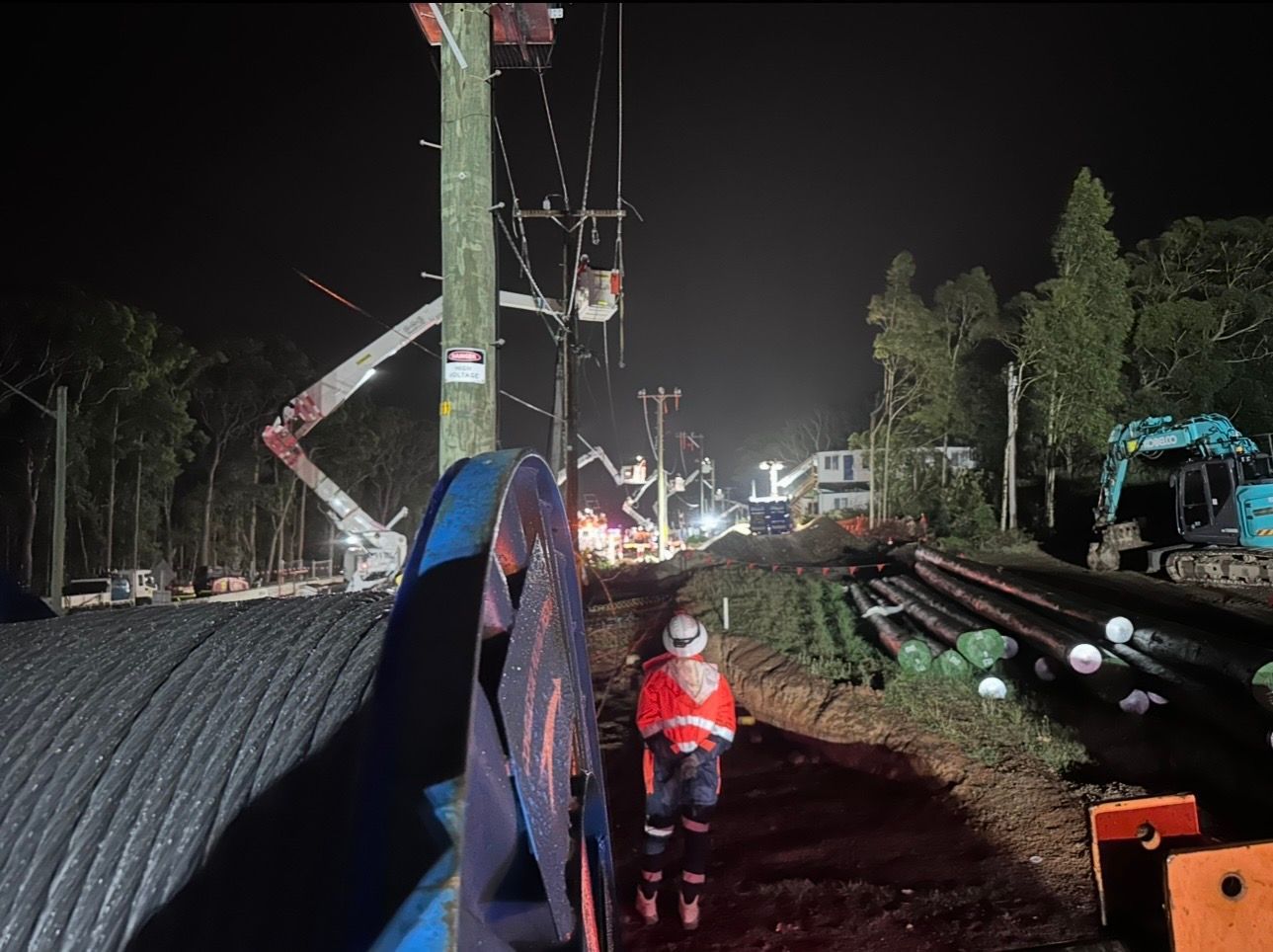 Nighttime railway construction site with a worker in a red jacket near tracks and utility poles