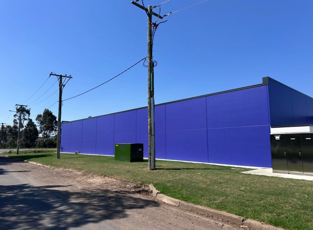 Blue industrial building beside a dirt road under a clear sky, with utility poles in front.
