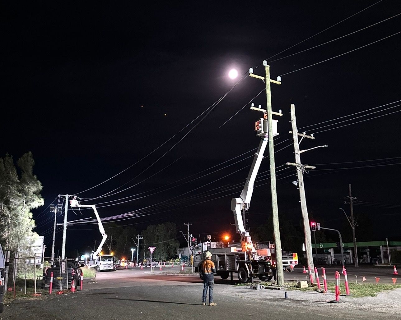 Night road work with utility poles, bucket trucks, workers, and overhead power lines under bright lights.