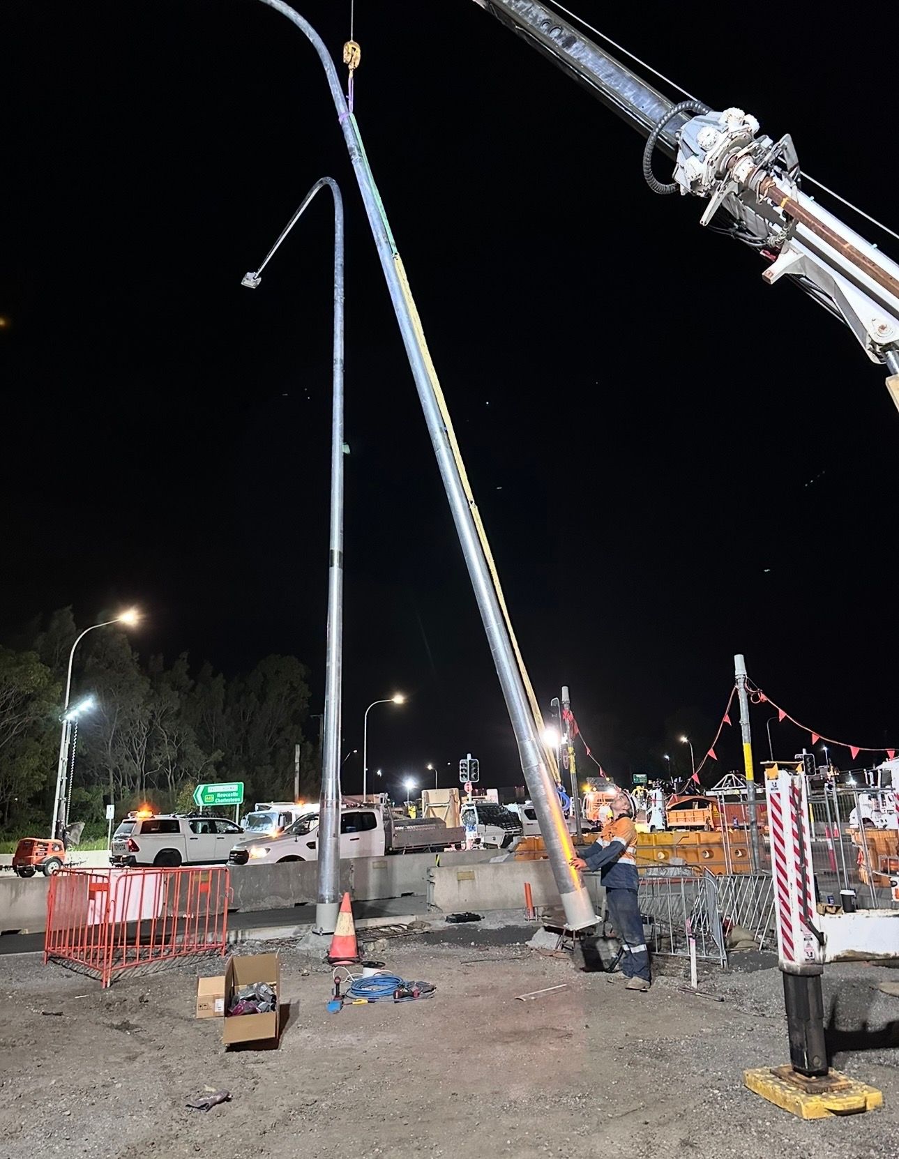 Night construction site with a crane lifting a light pole beside traffic barriers and work trucks