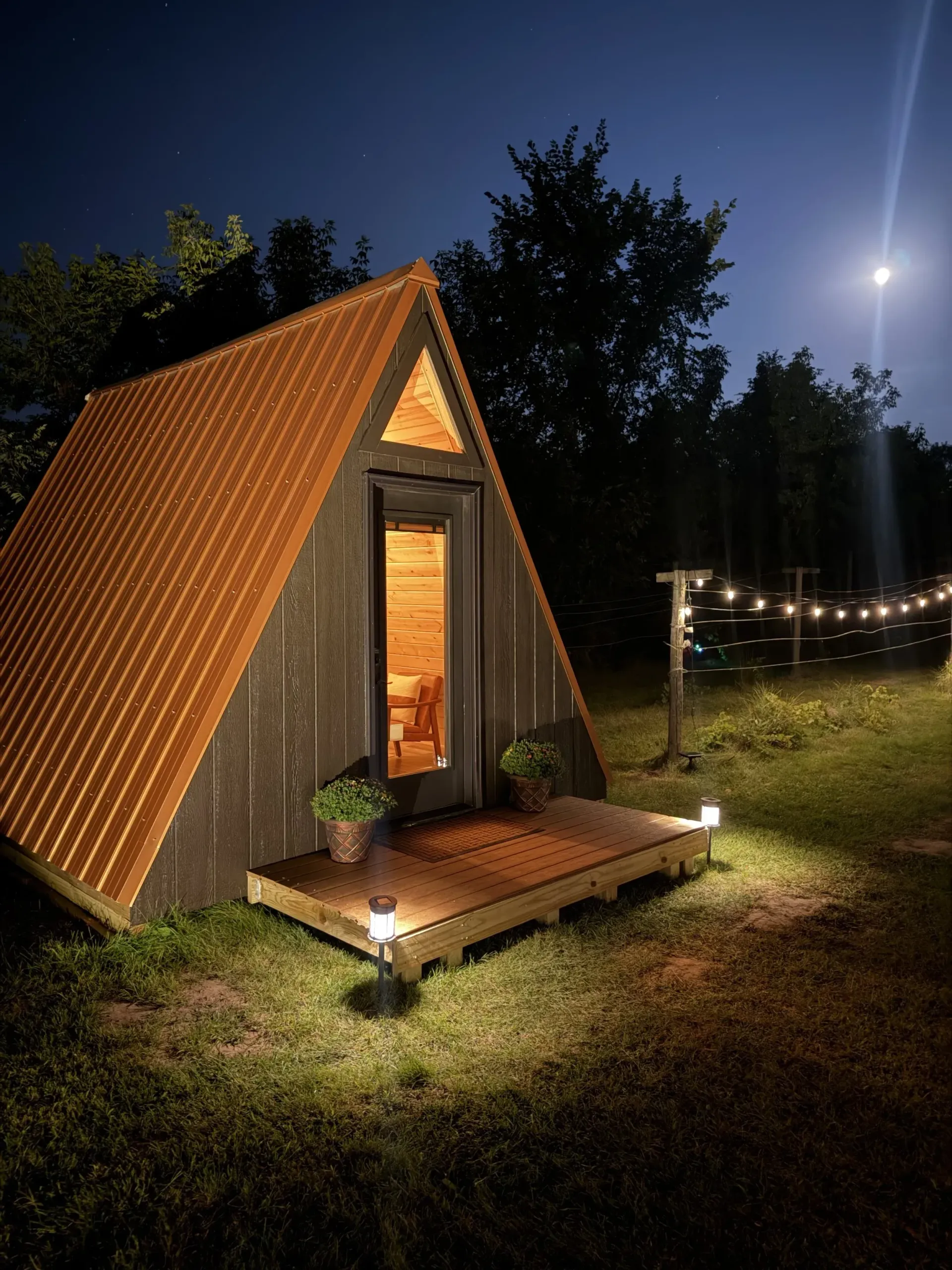 A small A-frame cabin glowing warmly at night, with string lights and a porch beside a moonlit grassy yard.
