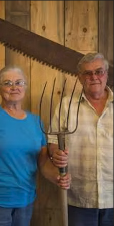 Al and Judy Olson standing indoors holding a pitchfork and a large saw against a wooden wall.