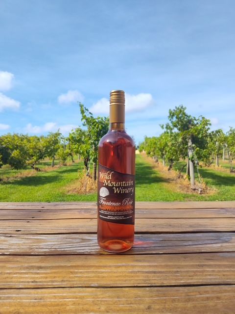 Bottle of amber liquor on a wooden table, with a grassy vineyard and blue sky in the background.