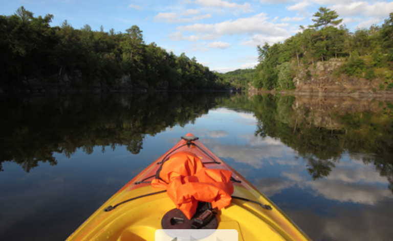 Yellow kayak gliding on a calm river, with trees reflecting on the water under a blue sky.