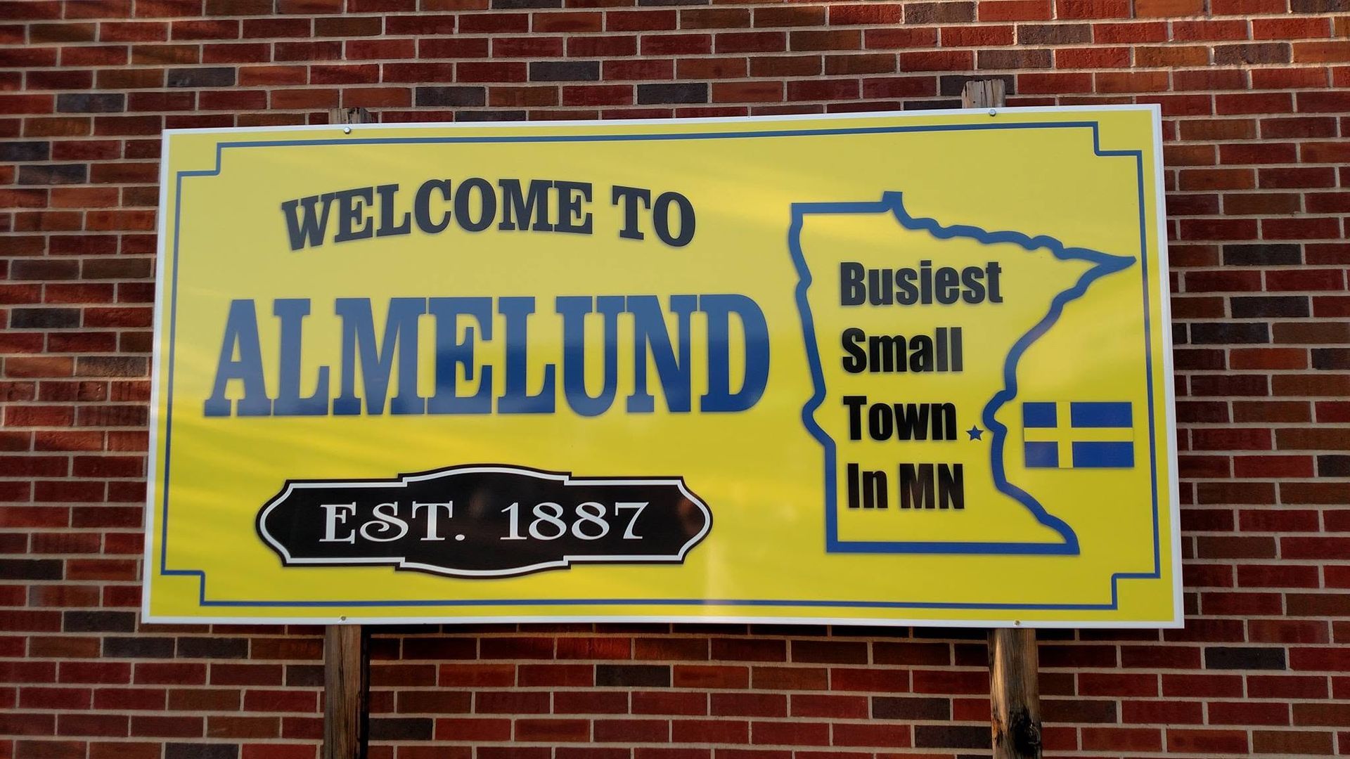 Yellow welcome sign for Almelund on a brick wall, reading “Welcome to Almelund” and “Smallest Town in MN.”