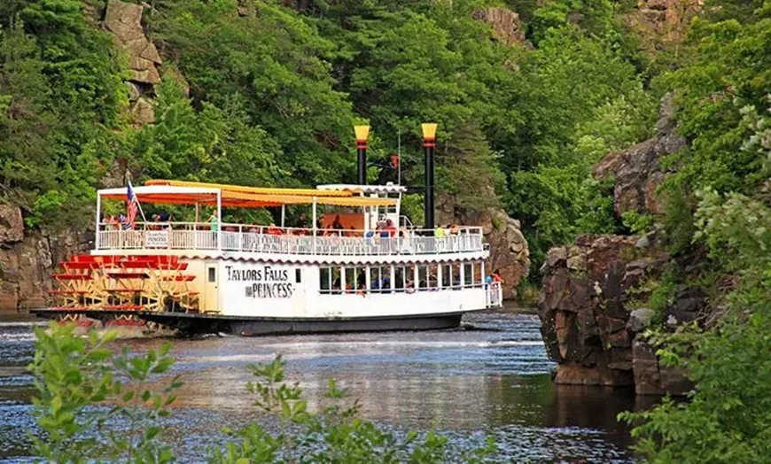 White sightseeing boat on a river beside rocky, forested cliffs under bright greenery