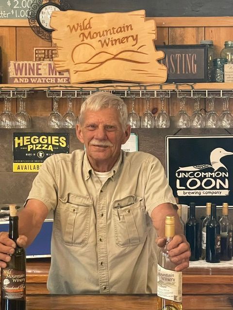 Man behind wine bar holding two wine bottles, with Wild Mountain Winery signs and hanging glasses in background