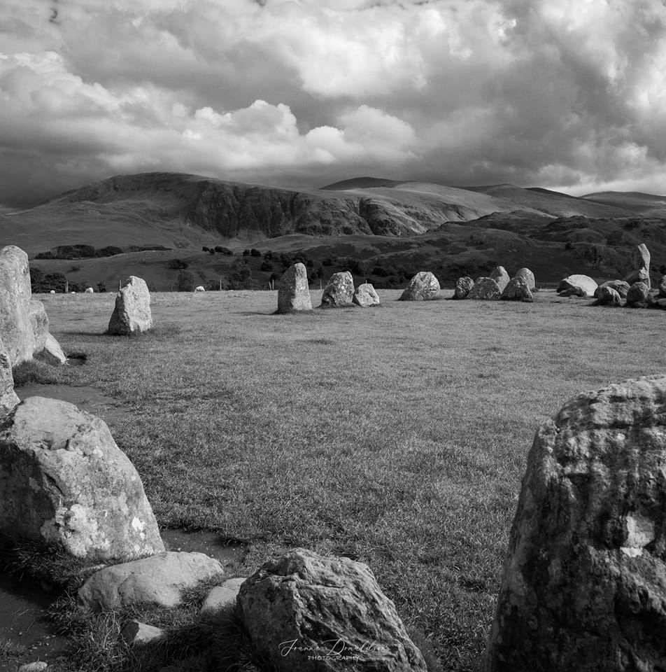 Stone circle on a grassy plain, mountains in the background under a cloudy sky.