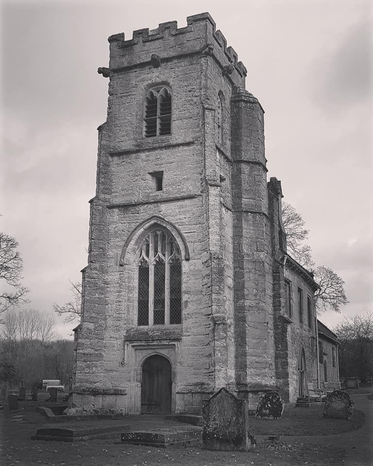 Black and white photo of an old stone church with a tall tower; it's a cloudy day in a graveyard.