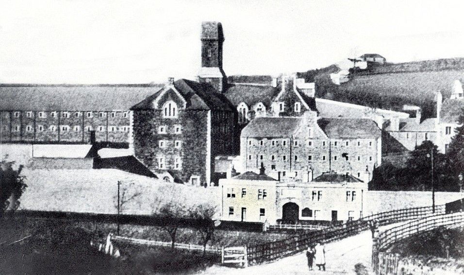 Historic black and white photo of a large, multi-building stone asylum with a prominent central tower, bridge, and hills.