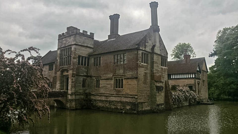 Moated medieval manor house on a cloudy day; stone walls, chimneys, water surrounding the building.
