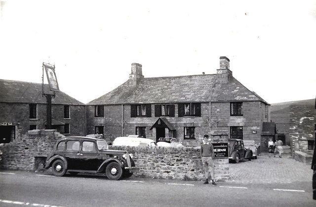 Black and white photo of a stone pub with parked cars in front. A sign hangs on a pole.