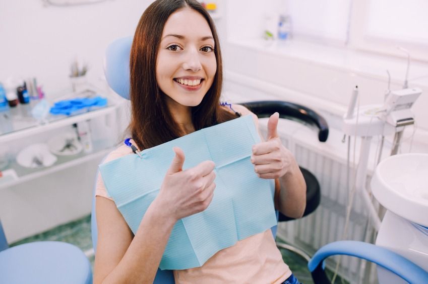 Smiling woman in a dentistโs office holding up a blue dental bib and giving thumbs up