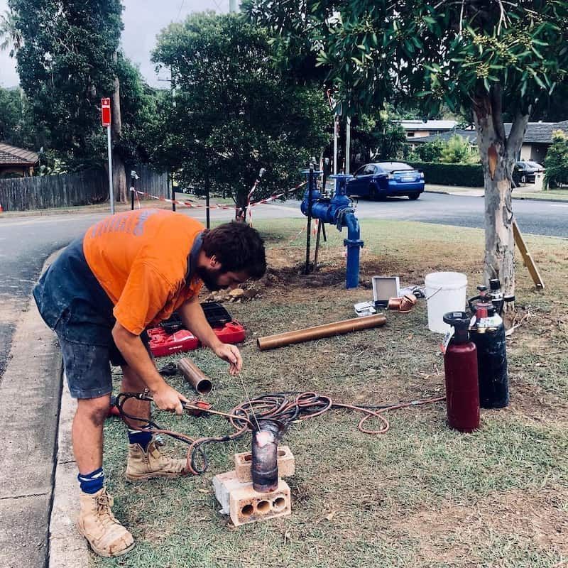 A Man in An Orange Shirt Is Working on A Pipe in The Grass — Nambucca Plumbing in Nambucca Heads, NSW
