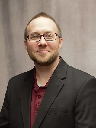 Man in glasses wearing a black blazer and maroon shirt, smiling against a gray backdrop