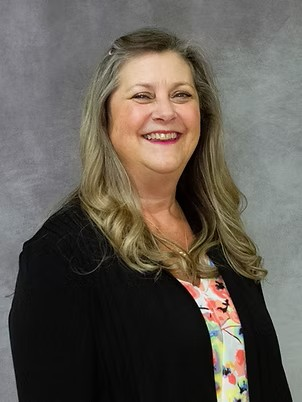 Smiling woman in a black blazer over a colorful blouse against a gray backdrop