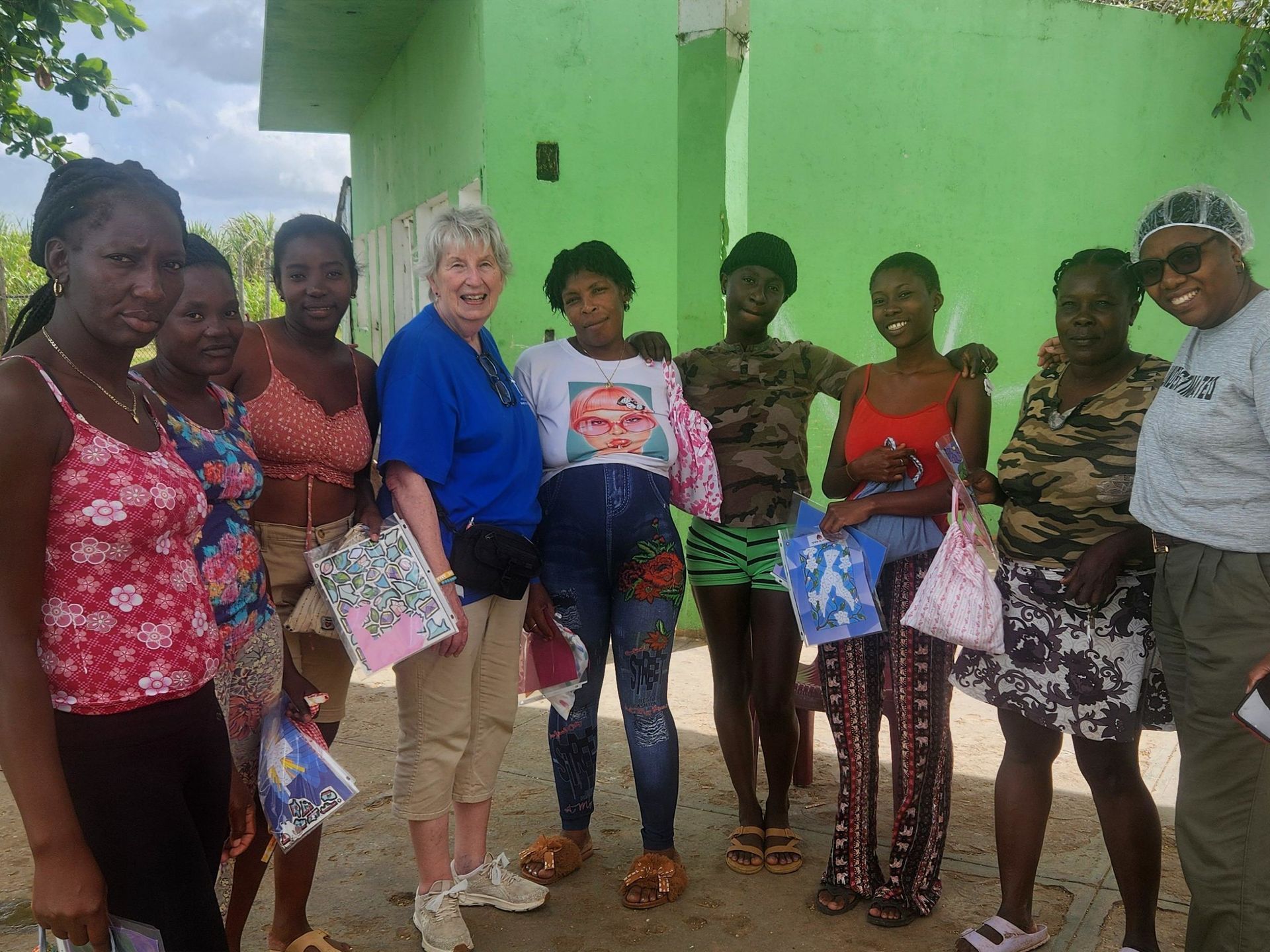 Group of people standing outdoors by a green building, holding bags and gifts, posing for a photo.