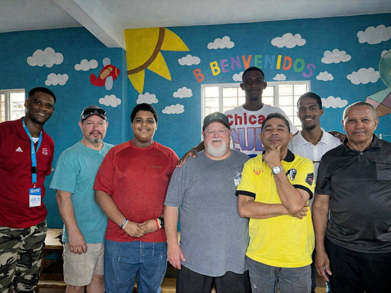 Group of people posing in front of a colorful “Bienvenidos” wall with cloud and sun mural.