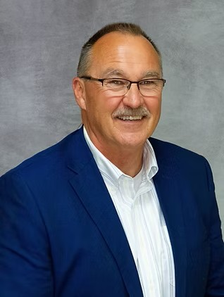 Man in blue blazer and white shirt smiling against a gray studio backdrop