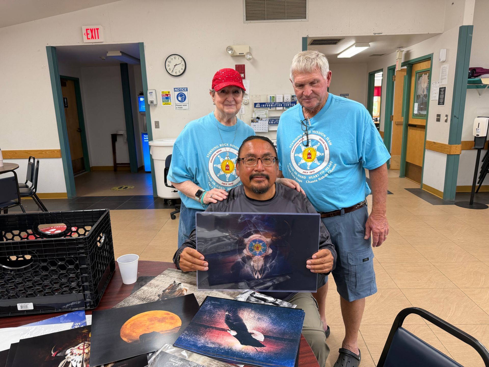 Three people in blue shirts pose indoors, holding cultural artwork at a table.