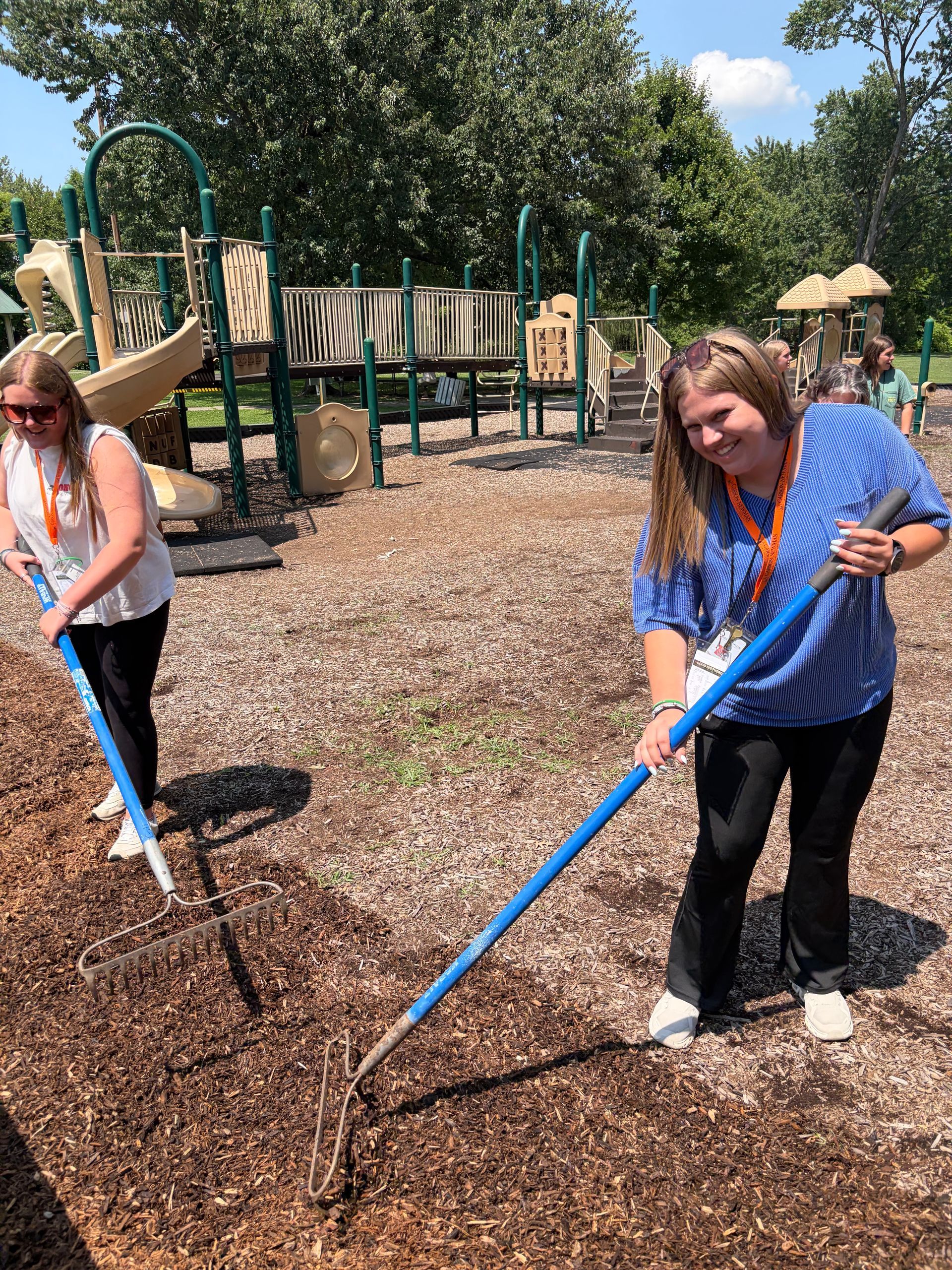 Two teenagers raking wood chips at a playground on a sunny day