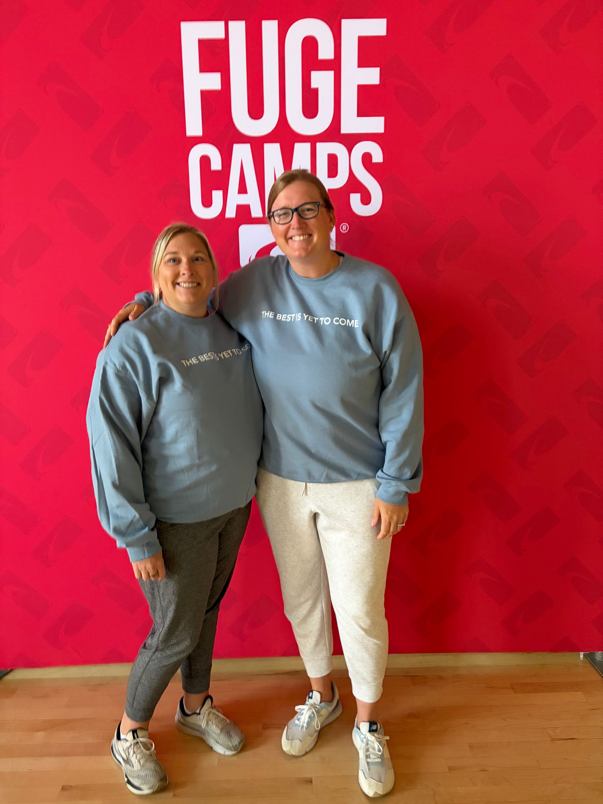 Two people in gray sweatshirts pose in front of a bright red FUGE CAMPS wall.