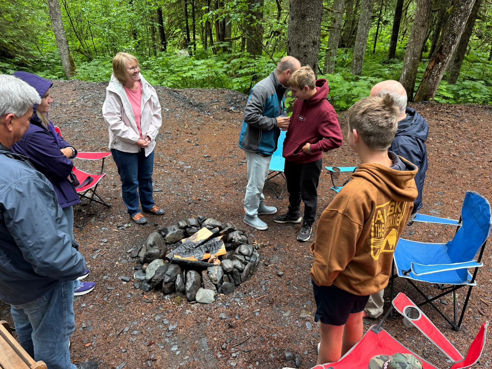 Group gathered around a small campfire in a forest clearing, with folding chairs and gravel ground.