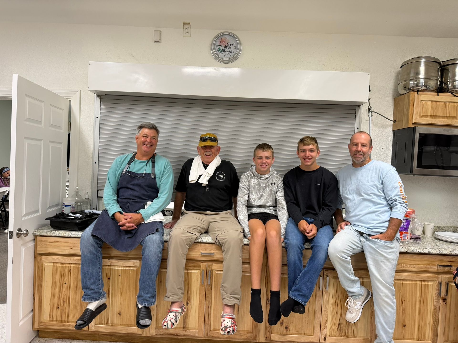 Five people sit on a kitchen counter, smiling in a bright home kitchen.