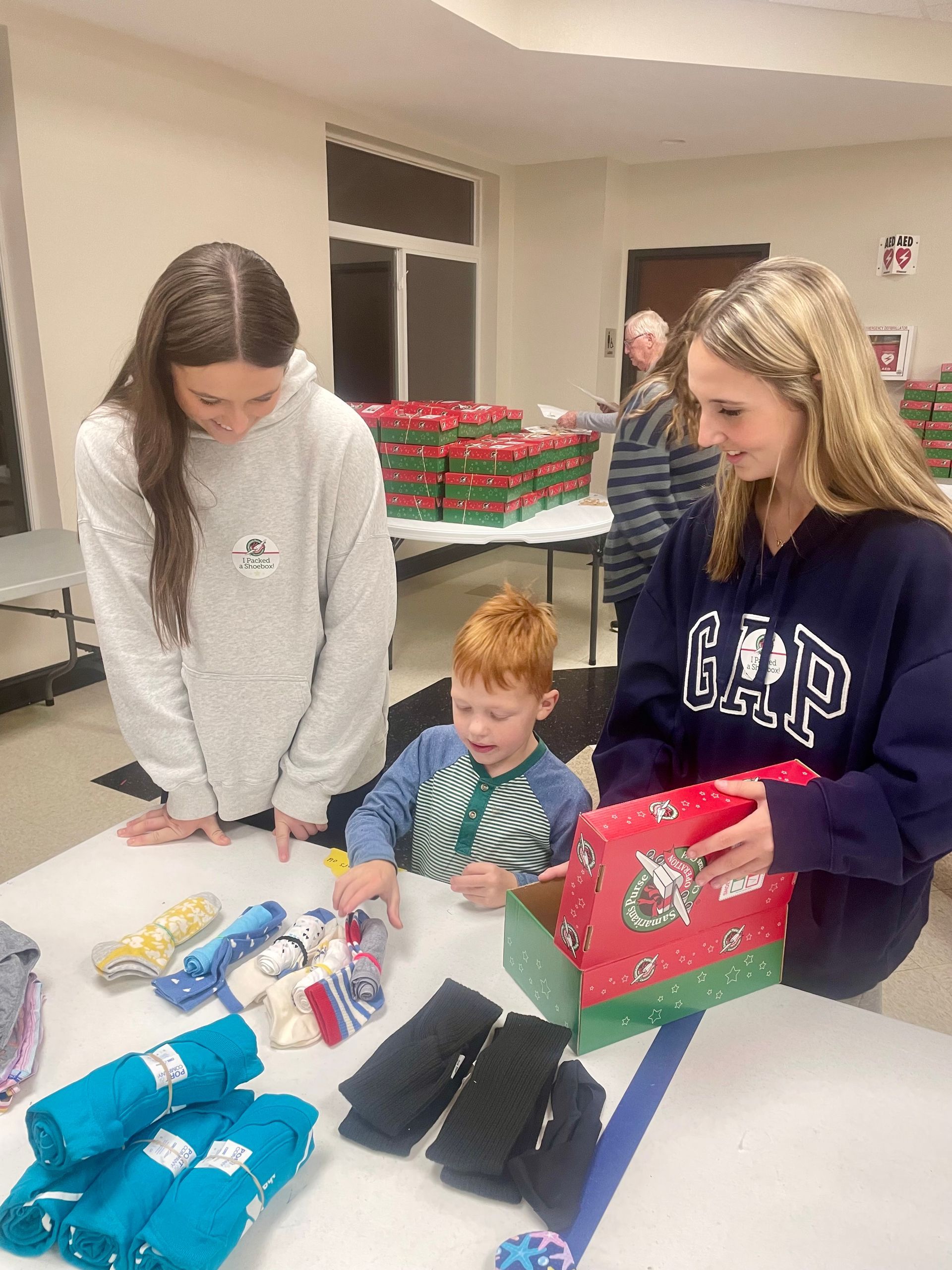 Three teenagers at a table sorting and wrapping colorful gift bags and items indoors