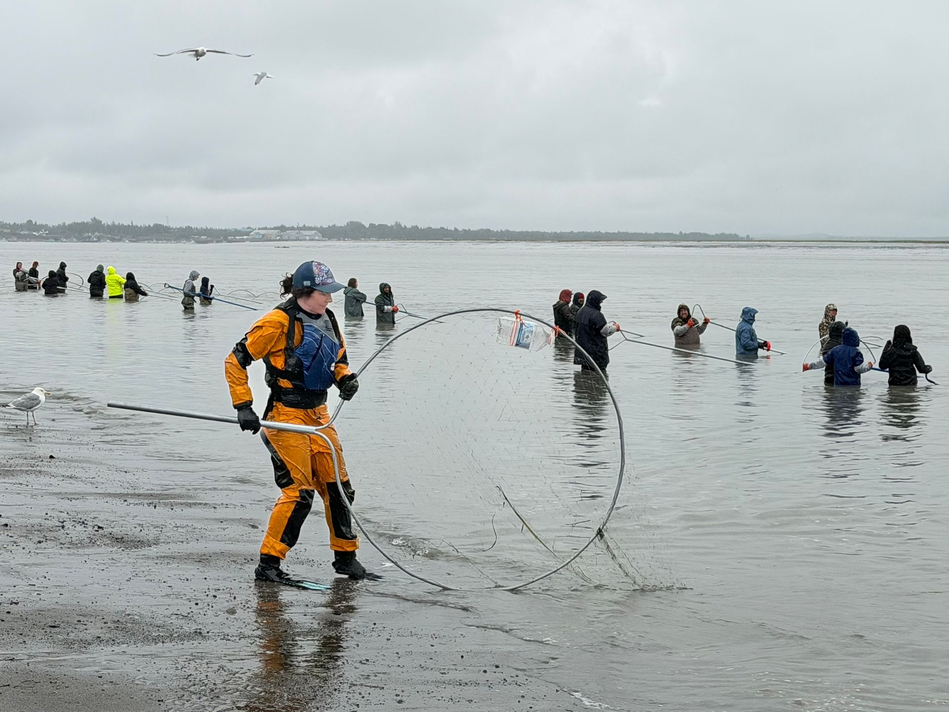 Person in orange rain gear fishing from a shallow shoreline with a group in the water under a gray sky