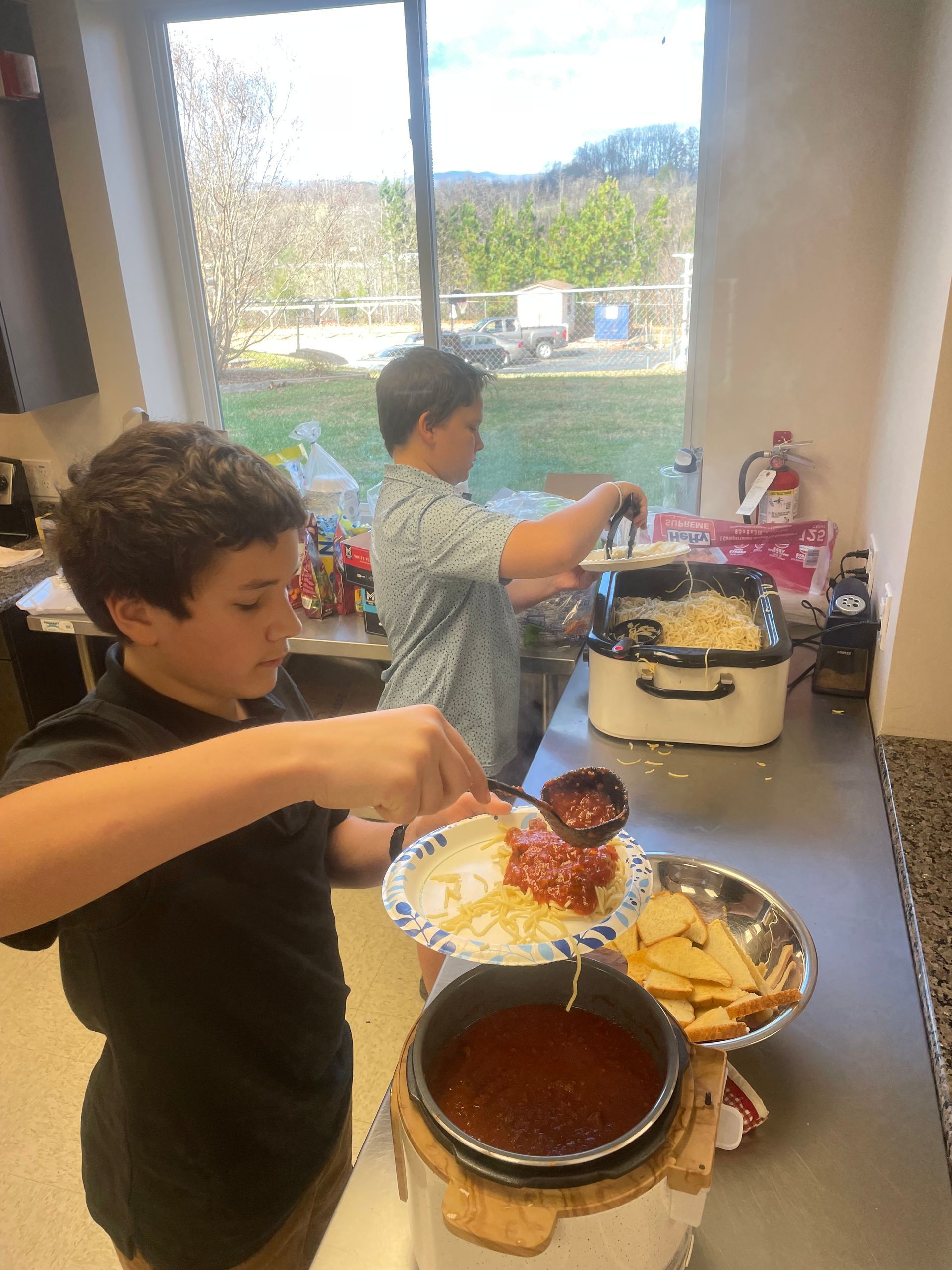 Two teenagers serving food at a kitchen counter with bowls, a pot, and a large window behind them