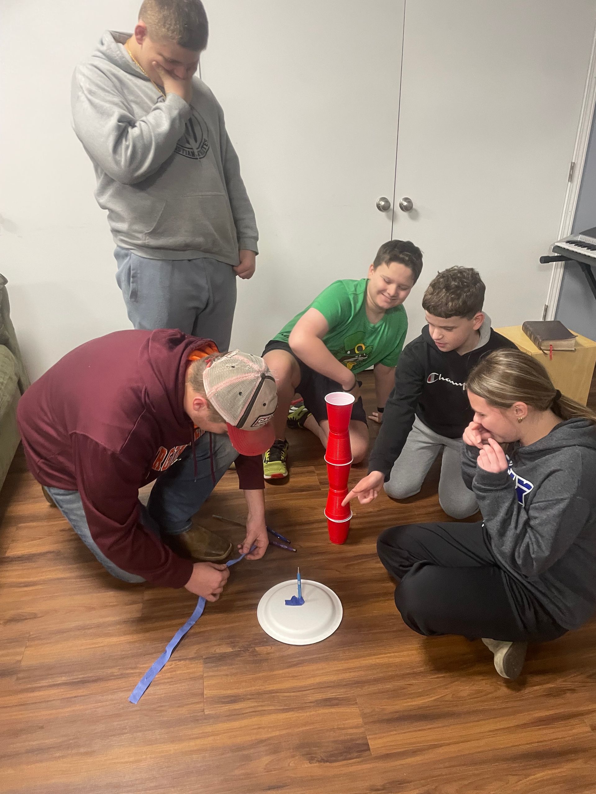 Five teenagers huddle around a red toy rocket on a wooden floor in a room.
