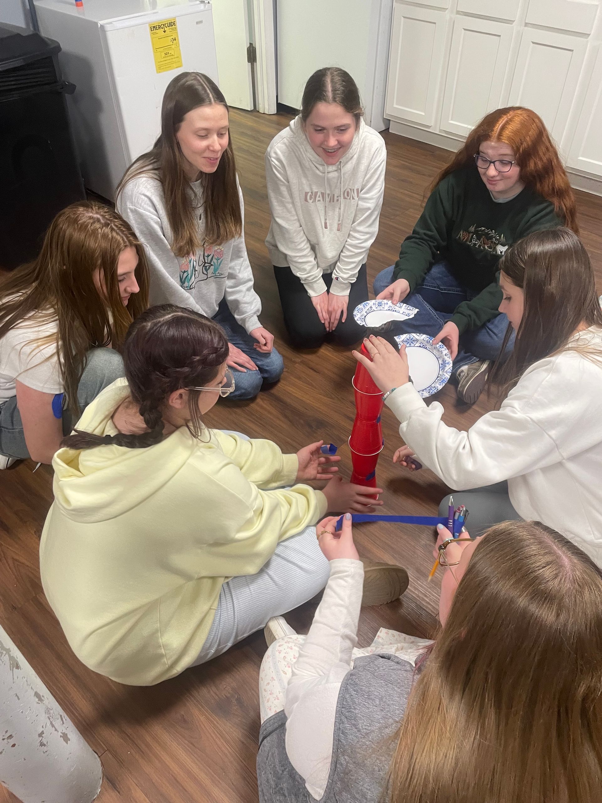 Group of teenagers sitting in a circle on the floor, collaborating around papers and a red bottle.