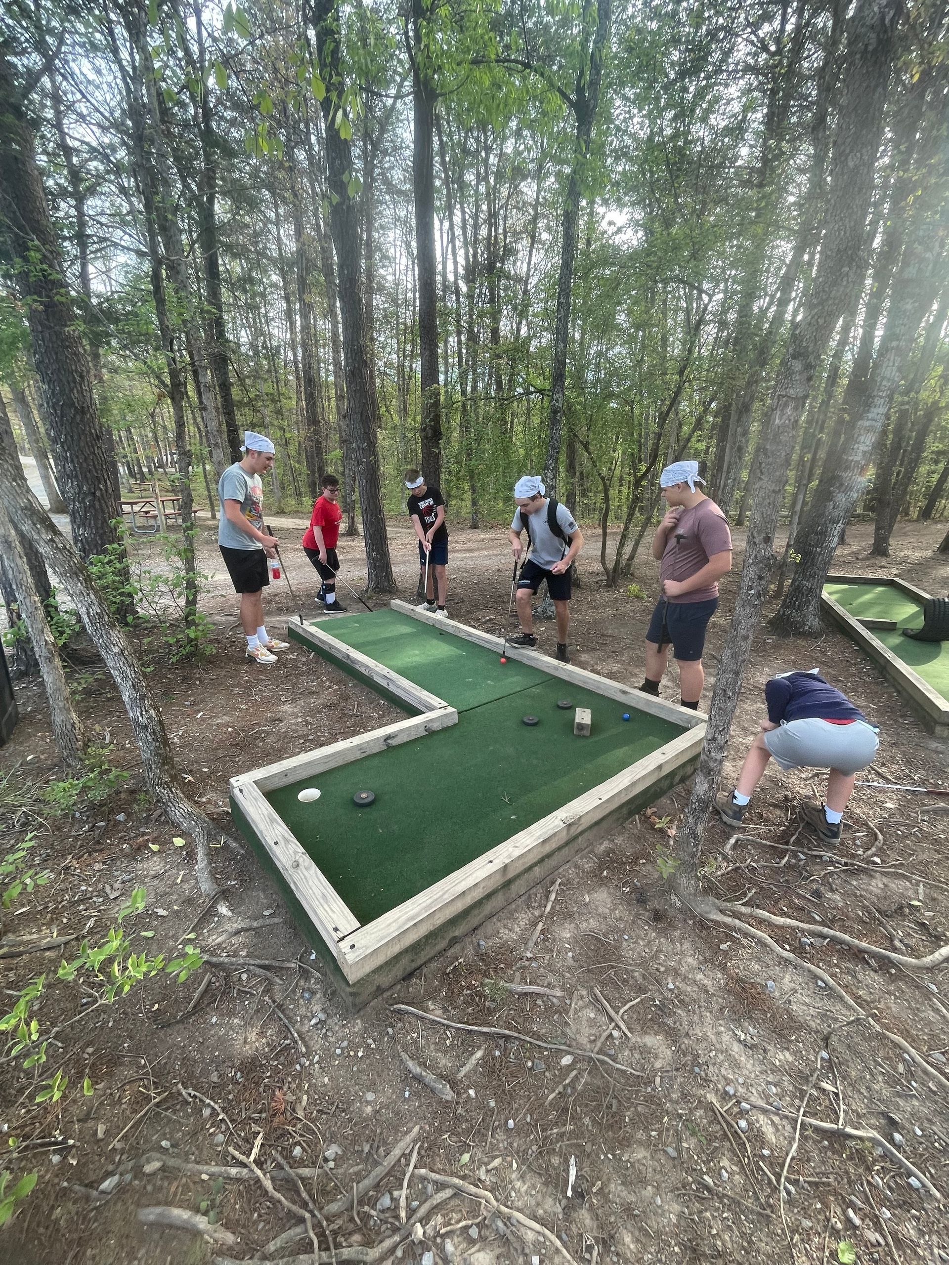 Teenagers playing mini golf on a forested course, with one person lining up a putt.