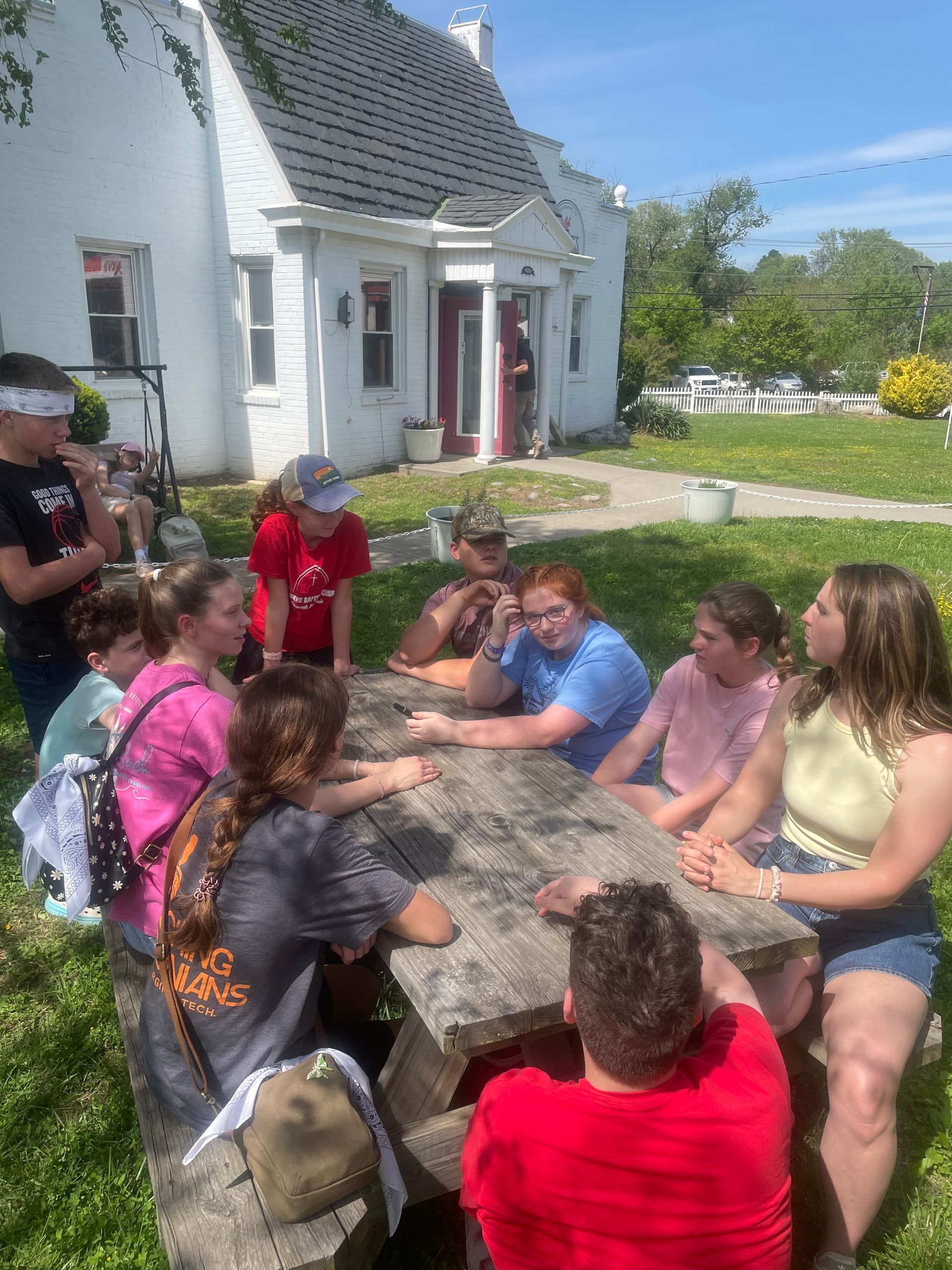 Teenagers sitting around a picnic table outside a white house on a sunny day