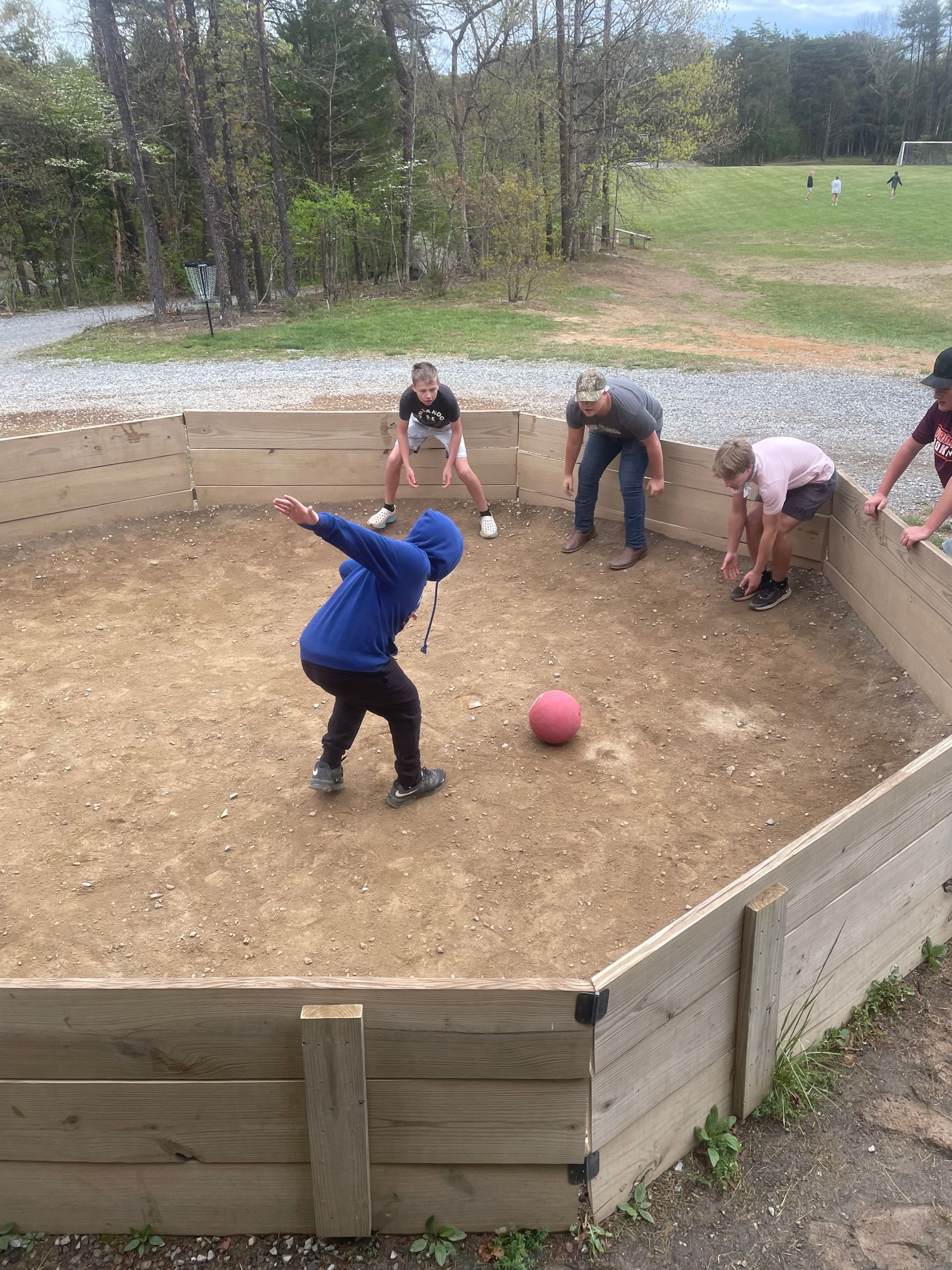 Teenagers playing bocce in a dirt court, with a red ball near the center.