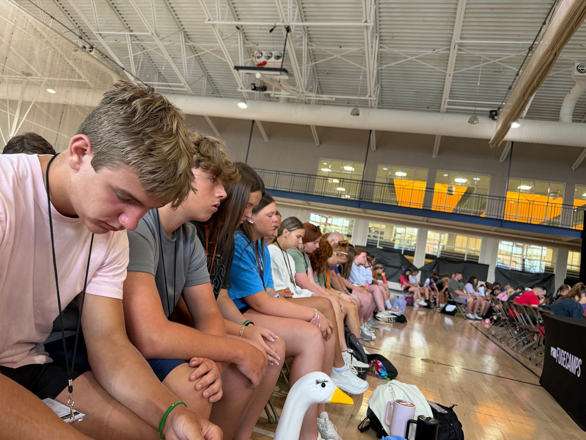Teenagers sitting along the edge of a gym floor watching an event, with ducks on the polished court.