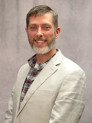 Smiling man in a light blazer and plaid shirt against a gray background