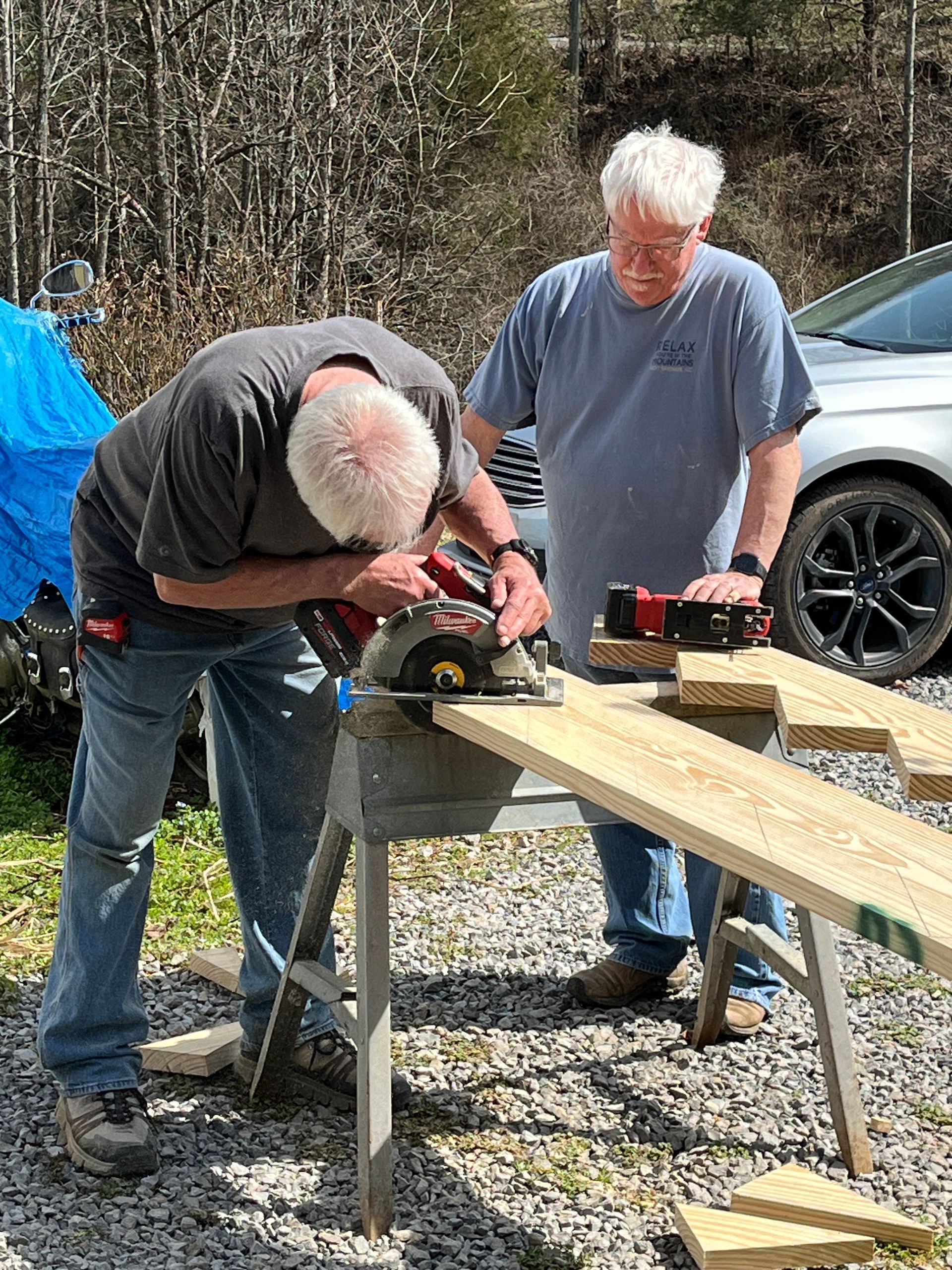 Two people sanding a wooden board on sawhorses outdoors near a car.