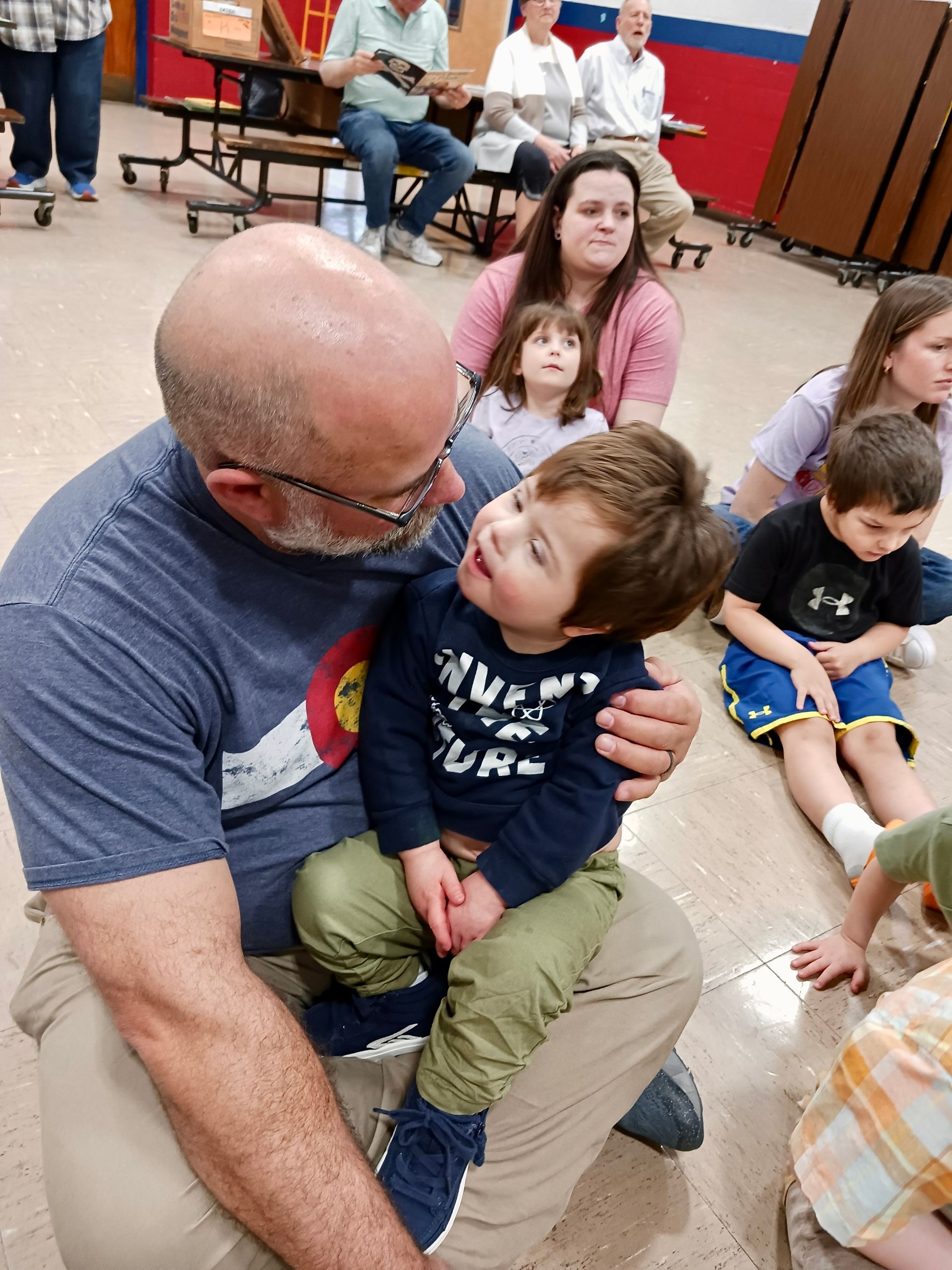 Man holding a smiling child on his lap in a gymnasium, with several children seated nearby.